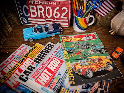 A stack of vintage magazines on a desk, with toy cars and a license plate in the background.