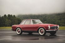 A red classic Mercedes-Benz parked in front of a lush foggy forest. 